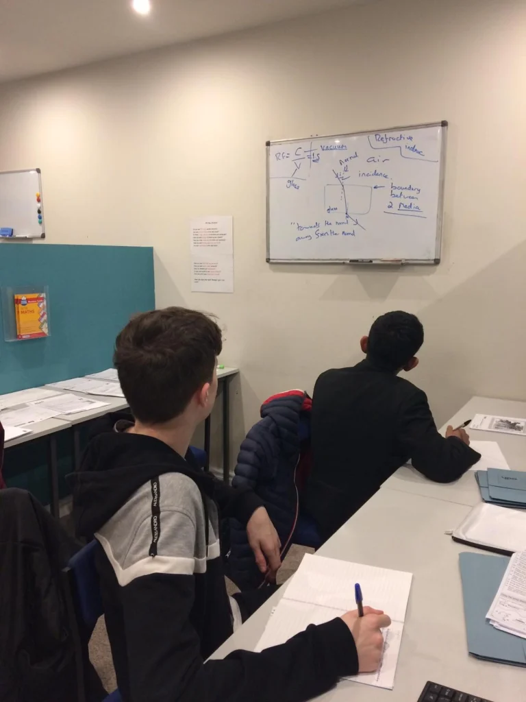 Two secondary students working at desks with a whiteboard behind them